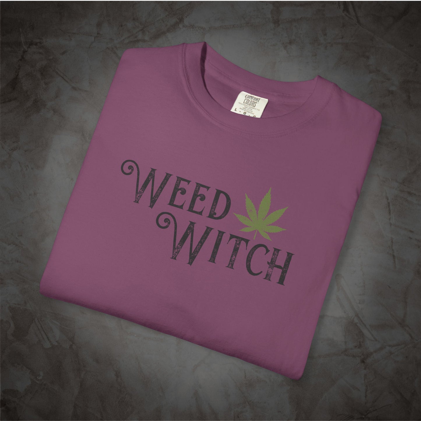 A folded purple t-shirt with a 'WEED WITCH' graphic in distressed black lettering and a green cannabis leaf, displayed on a dark textured background.