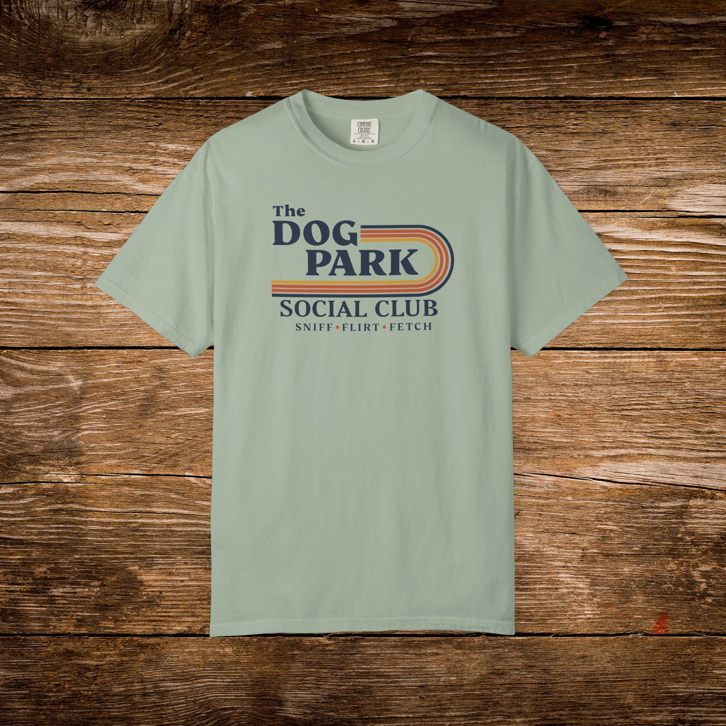 A sage green short-sleeve t-shirt with a retro-style graphic reading 'The DOG PARK SOCIAL CLUB' and 'SNIFF - FLIRT - FETCH' in navy, orange, and yellow, displayed flat against a rustic brown wooden background. The design text reads "Sniff, Flirt, Fetch."