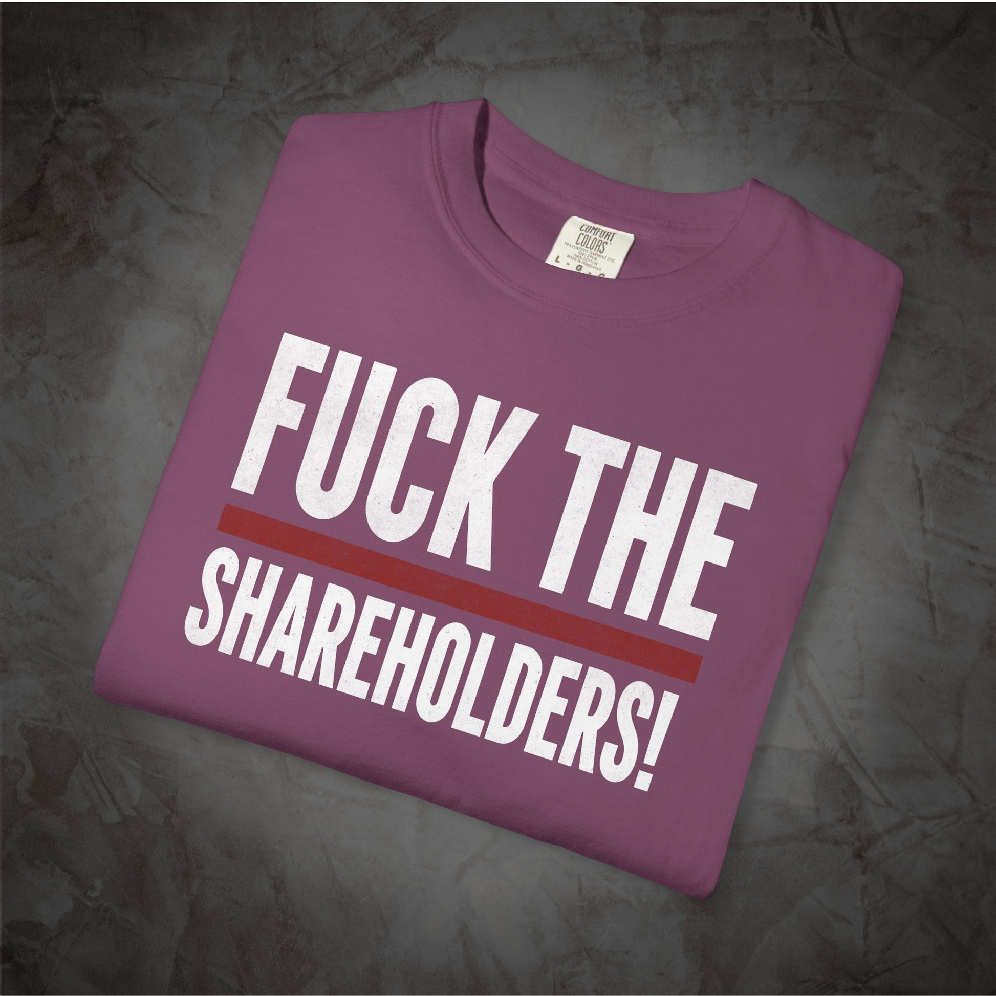 A folded purple t-shirt with the text 'FUCK THE SHAREHOLDERS!' printed in white, separated by a red horizontal stripe, displayed on a dark textured background.