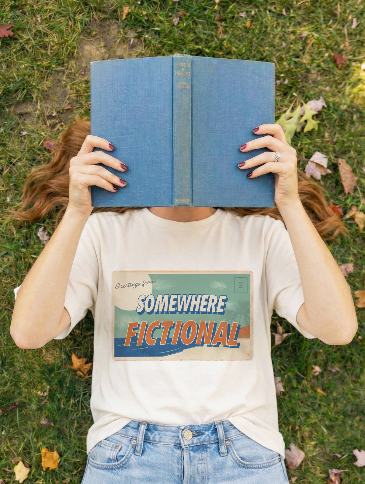 Person wearing an ivory “Greetings from Somewhere Fictional” graphic t-shirt while holding a blue book in front of their face outdoors.