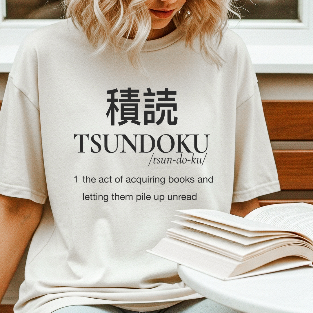 Person reading while wearing an ivory Tsundoku dictionary definition T-shirt beside a stack of books.