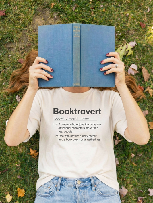 Person holding a blue hardcover book while wearing a Booktrovert dictionary definition T-shirt outdoors.