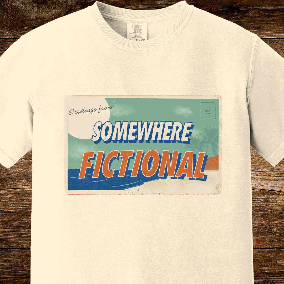 Ivory Comfort Colors t-shirt featuring a vintage-inspired “Greetings from Somewhere Fictional” postcard graphic in teal, cream, and orange.