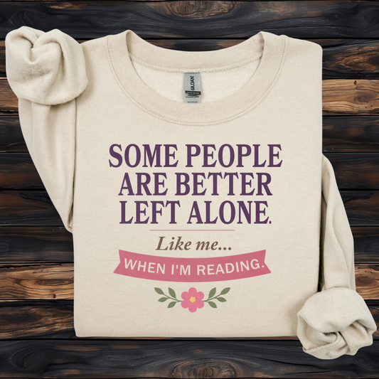 A cream-colored long-sleeve t-shirt with rolled cuffs, displayed flat on a dark wood background. The shirt features a graphic print in purple and pink text that reads 'SOME PEOPLE ARE BETTER LEFT ALONE.