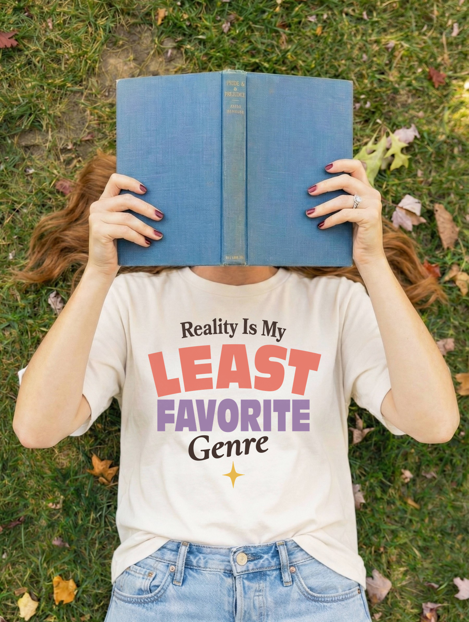 A person lying on grass outdoors holds a blue book over their face, wearing a white graphic T-shirt that reads 'Reality Is My LEAST FAVORITE Genre' in red, purple, and black text, with light blue jeans.