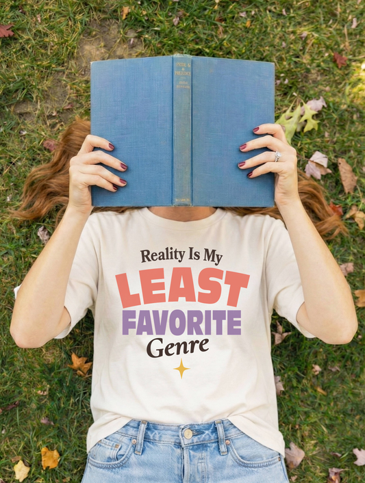 A person lying on grass outdoors holds a blue book over their face, wearing a white graphic T-shirt that reads 'Reality Is My LEAST FAVORITE Genre' in red, purple, and black text, with light blue jeans.
