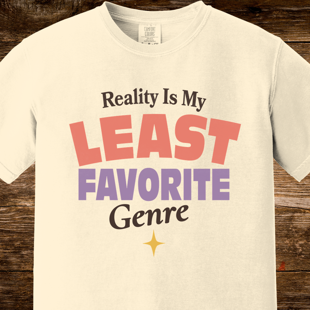 An off-white crewneck t-shirt with a retro-style graphic print reading 'Reality Is My LEAST FAVORITE Genre' in black, coral pink, and purple lettering, displayed on a rustic dark wood background.