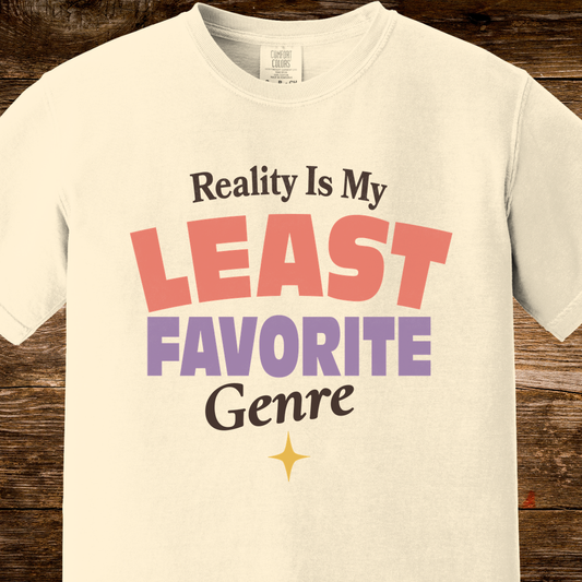 An off-white crewneck t-shirt with a retro-style graphic print reading 'Reality Is My LEAST FAVORITE Genre' in black, coral pink, and purple lettering, displayed on a rustic dark wood background.