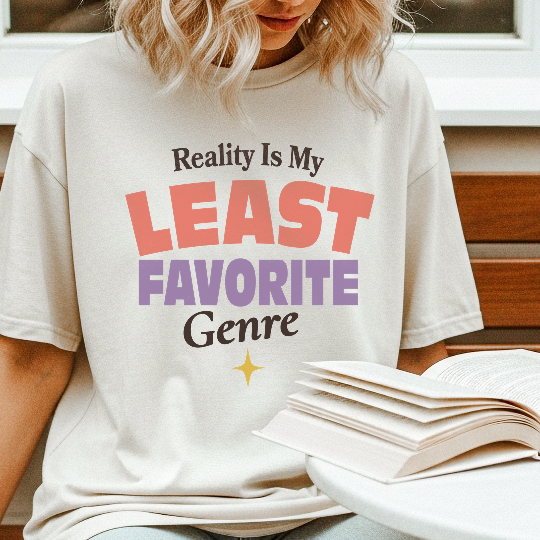A woman wearing a cream-colored graphic t-shirt with the text 'Reality Is My LEAST FAVORITE Genre' in coral, purple, and black lettering. She is sitting next to an open book on a white table.