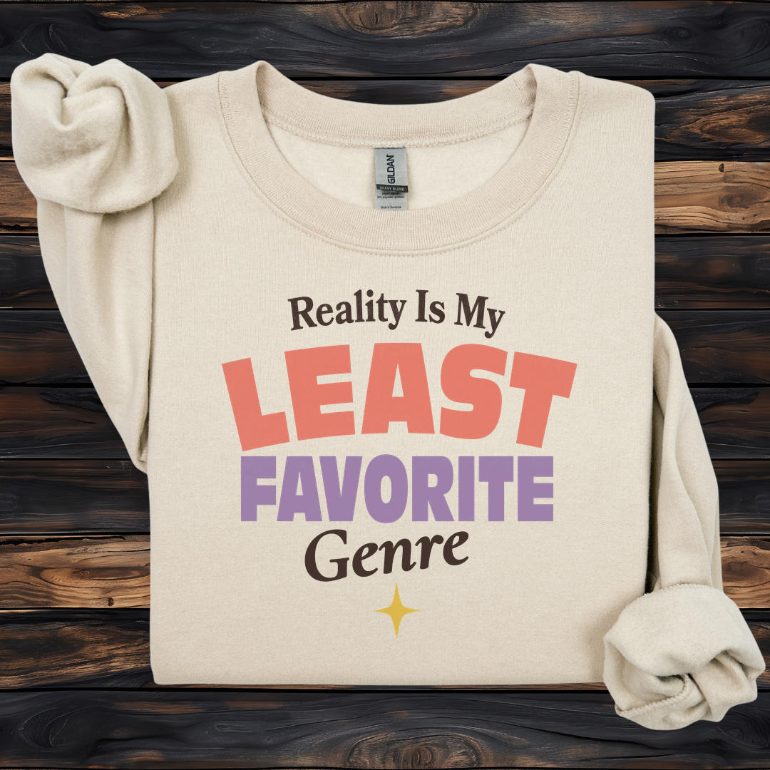 A folded cream-colored long-sleeve t-shirt with rolled cuffs, featuring a graphic print that reads 'Reality Is My LEAST FAVORITE Genre' in black, coral pink, and purple lettering, displayed on a dark wood background.