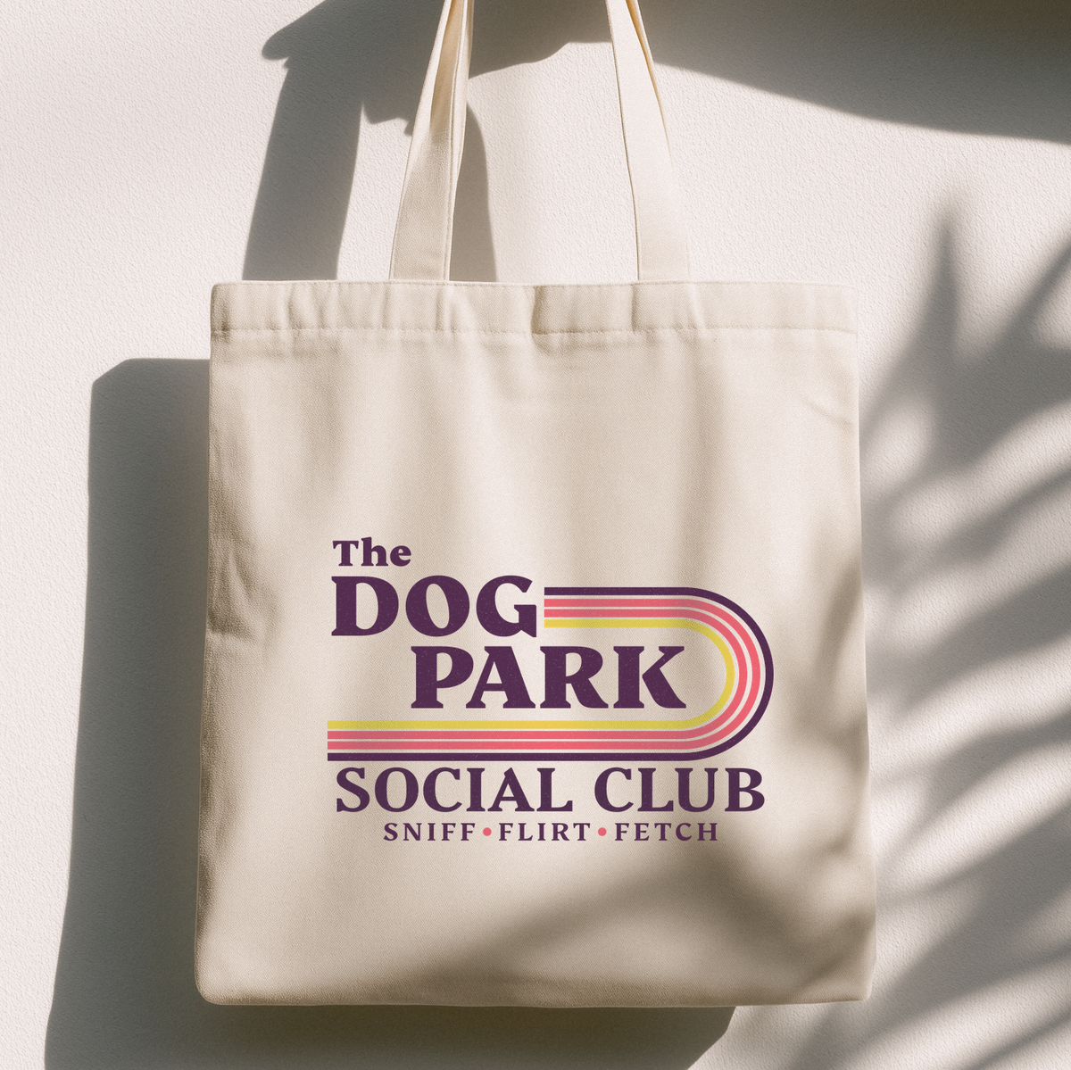 An off-white canvas tote bag with a colorful retro-style graphic that reads 'The DOG PARK SOCIAL CLUB' and 'SNIFF • FLIRT • FETCH'. The bag is hanging against a white wall with soft shadow patterns cast across it.