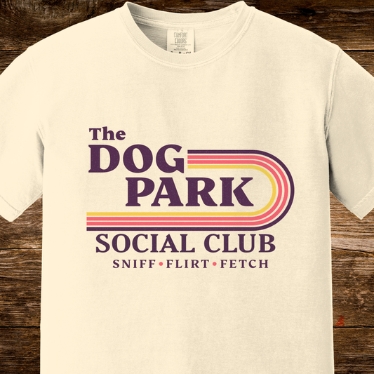 An off-white crewneck t-shirt featuring a retro-style graphic that reads 'The DOG PARK SOCIAL CLUB' with 'SNIFF • FLIRT • FETCH' underneath. The graphic includes purple text and yellow, orange, and purple retro stripes.