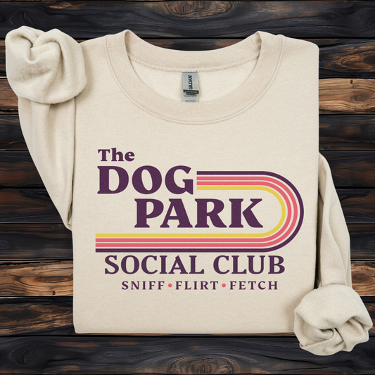 A folded cream-colored long-sleeve t-shirt with rolled cuffs, featuring a retro-style graphic that reads 'The DOG PARK SOCIAL CLUB' and 'SNIFF • FLIRT • FETCH' in purple, pink, and yellow. The shirt is displayed flat on a dark wood plank background.