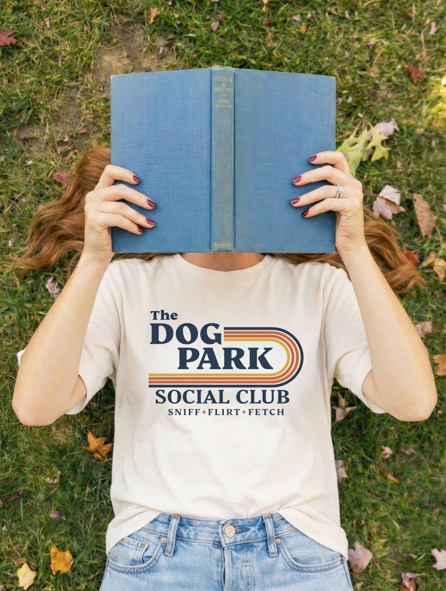 A person lying on grass outdoors holds a blue book over their face, wearing a cream graphic T-shirt that reads 'The Dog Park Social Club' with a retro stripe design and light blue jeans. The design text reads "Sniff, Flirt, Fetch."