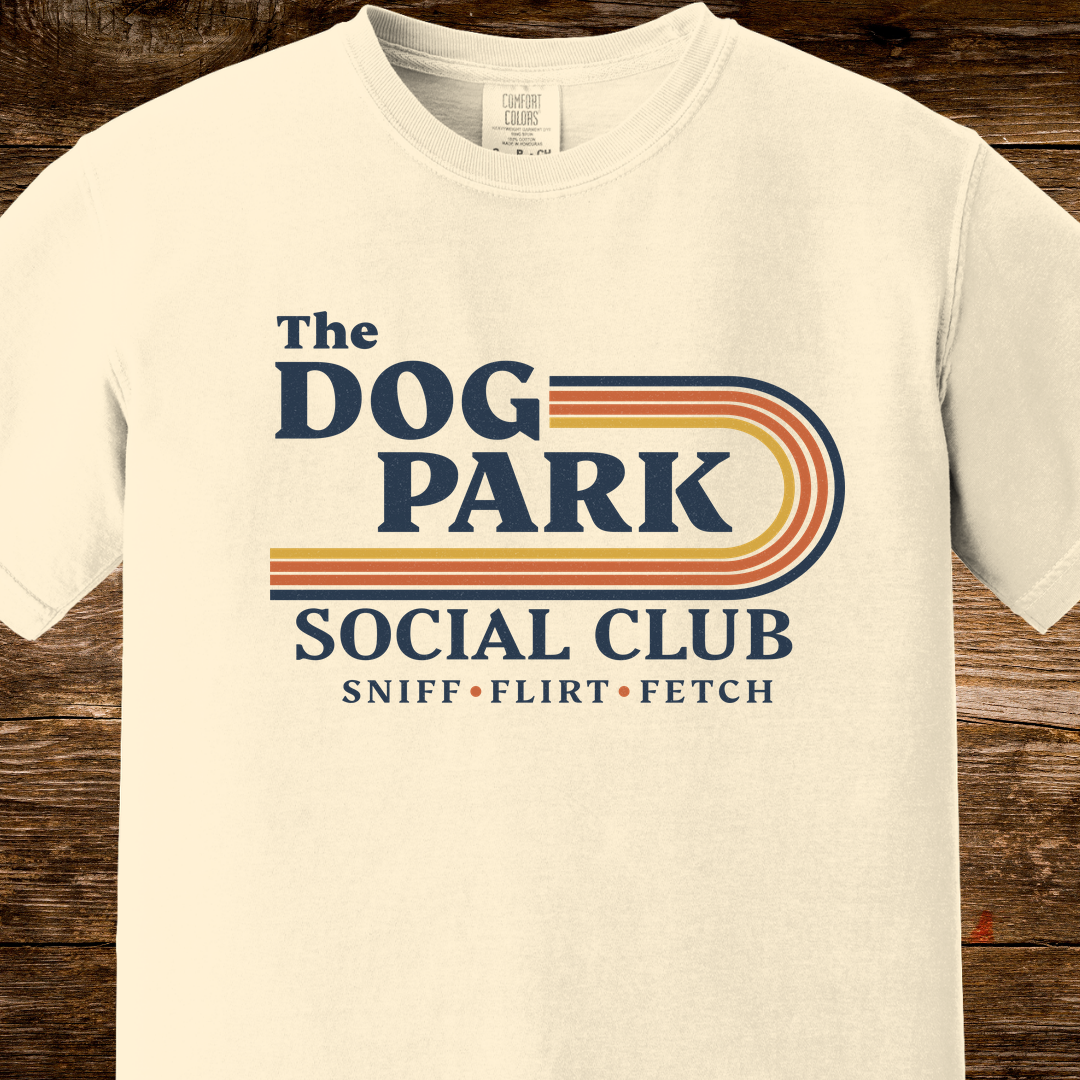 An off-white crewneck t-shirt featuring a retro-style graphic that reads 'The DOG PARK SOCIAL CLUB' with 'SNIFF • FLIRT • FETCH' underneath, displayed on a rustic dark wood background. The design text reads "Sniff, Flirt, Fetch."