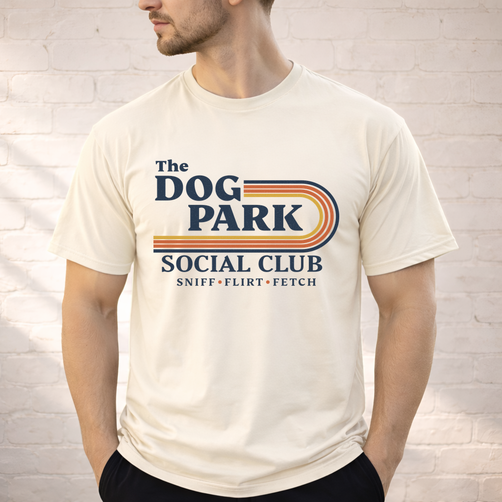 A man wearing a cream-colored t-shirt with a retro-style graphic that reads 'The DOG PARK SOCIAL CLUB' and 'SNIFF • FLIRT • FETCH' in navy blue and orange and yellow stripes, standing against a white brick wall. The design text reads "Sniff, Flirt, Fetch."
