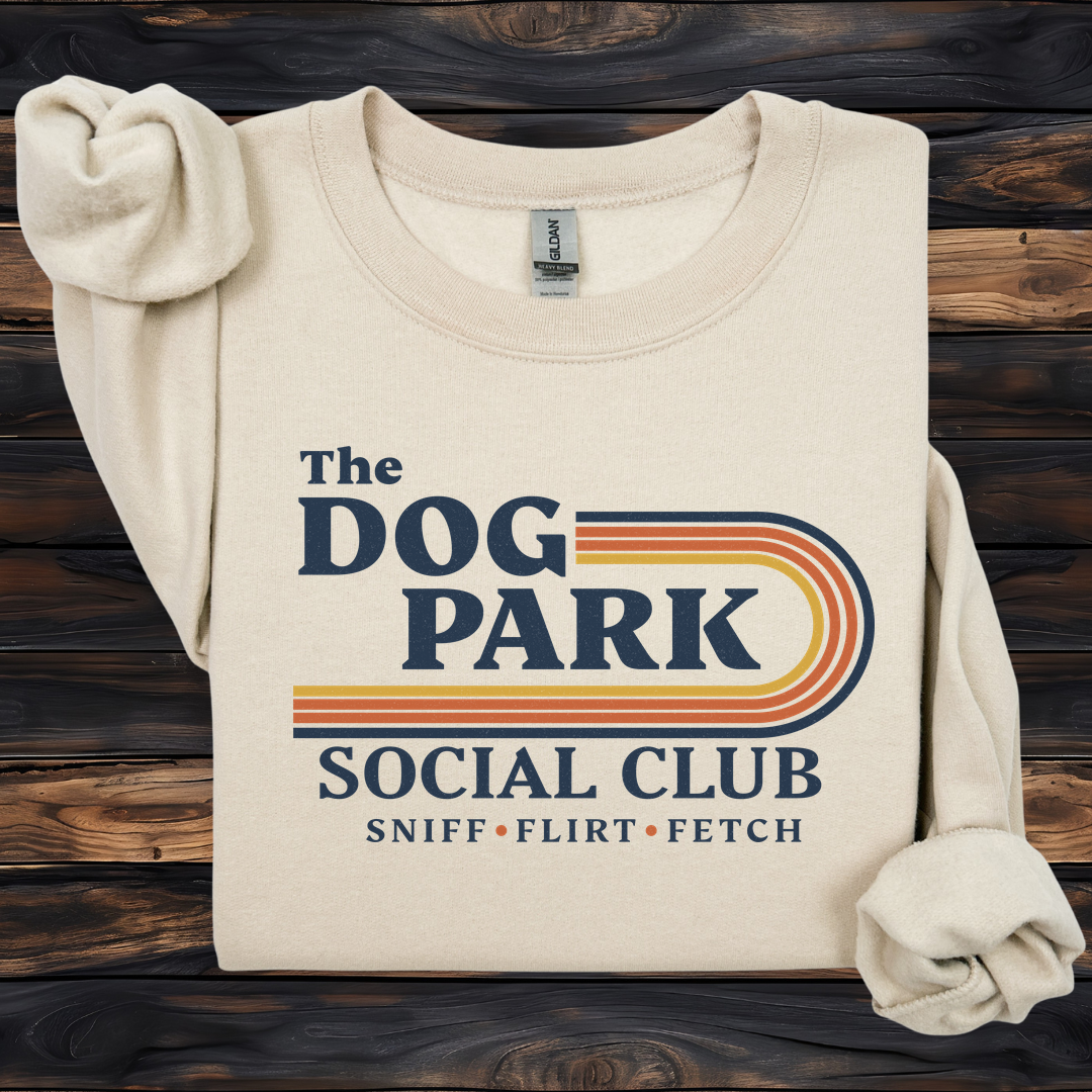 A folded cream-colored sweatshirt with rolled sleeves, featuring a retro-style graphic that reads 'The DOG PARK SOCIAL CLUB' and 'SNIFF • FLIRT • FETCH' in navy and orange-yellow stripes, displayed on a dark wood plank background. The design text reads "Sniff, Flirt, Fetch."