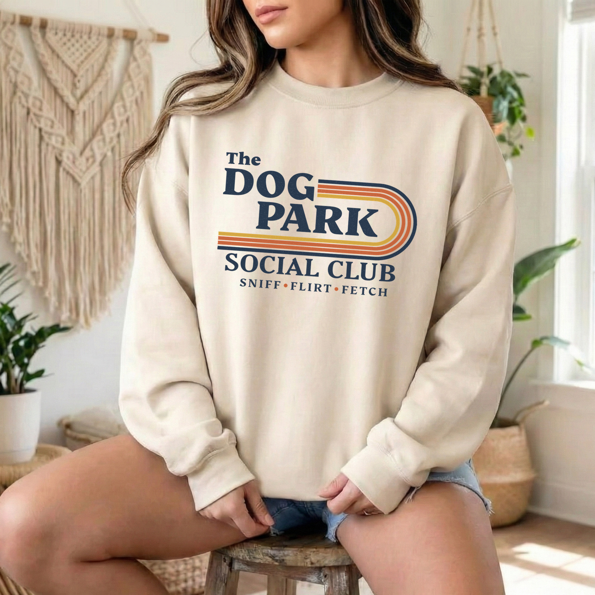 A woman wearing a cream-colored crewneck sweatshirt with a retro-style 'The Dog Park Social Club' graphic, sitting on a wooden stool in a cozy, boho-style room. The design text reads "Sniff, Flirt, Fetch."