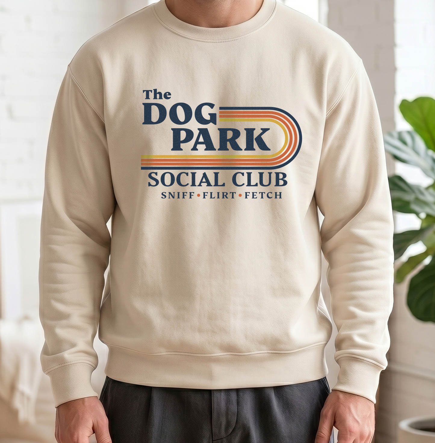 A person wearing a cream-colored crewneck sweatshirt with a retro-style graphic that reads 'The DOG PARK SOCIAL CLUB' and 'SNIFF • FLIRT • FETCH' in navy and orange-yellow tones. The background is a bright, softly blurred indoor setting with a white brick wall and a green plant. The design text reads "Sniff, Flirt, Fetch."