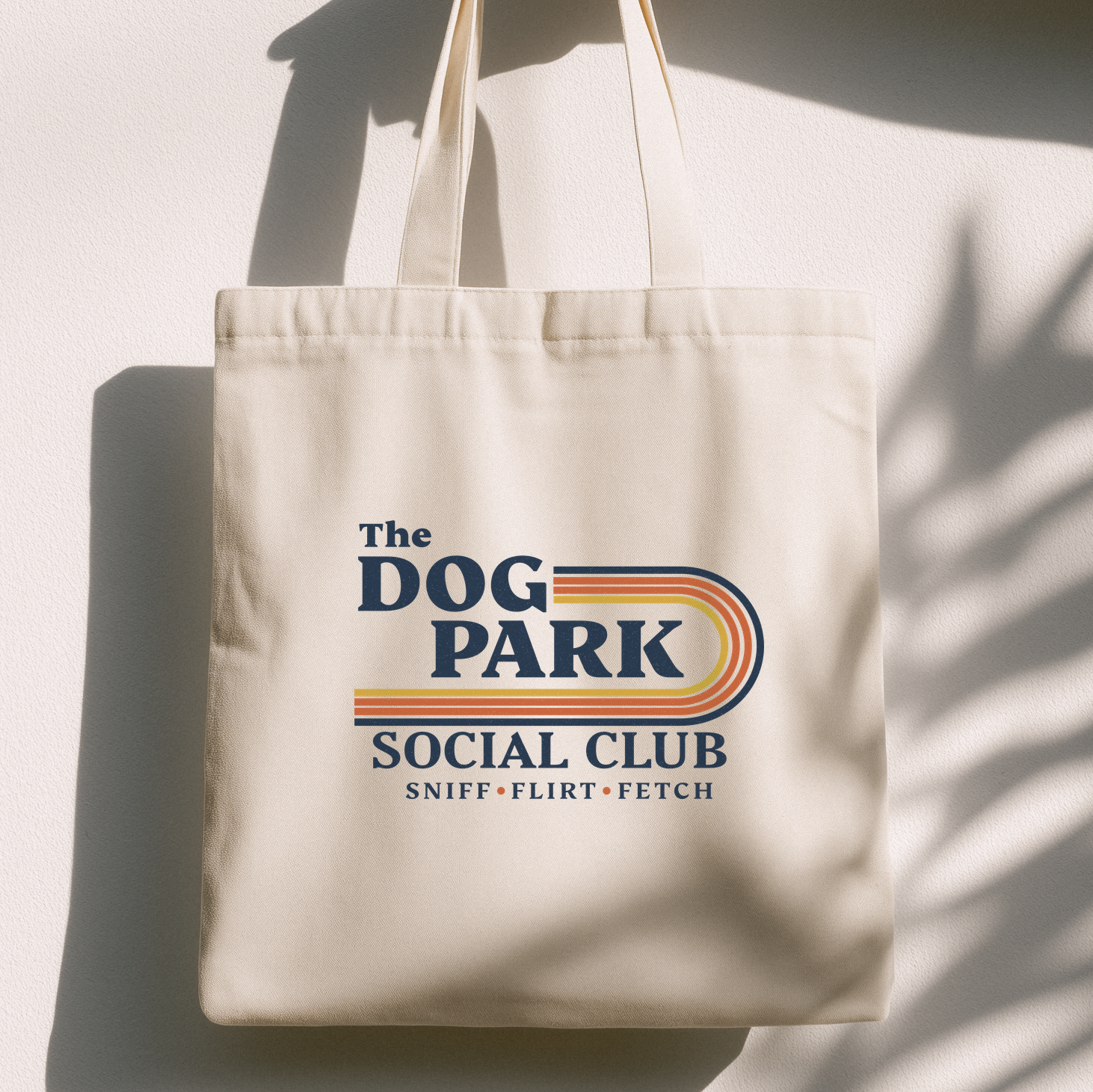 An off-white canvas tote bag with a retro-style graphic reading 'The DOG PARK SOCIAL CLUB' and smaller text 'SNIFF • FLIRT • FETCH'. The bag is photographed hanging against a white wall with strong sunlight and leaf shadows cast across it. The design text reads "Sniff, Flirt, Fetch."