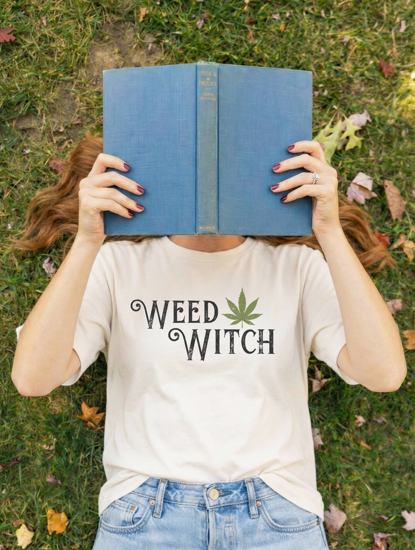 A person lying on grass outdoors holds a blue book over their face. They are wearing a white graphic T-shirt with the text 'READ BOOKS 2028' in navy and red, paired with light blue jeans.