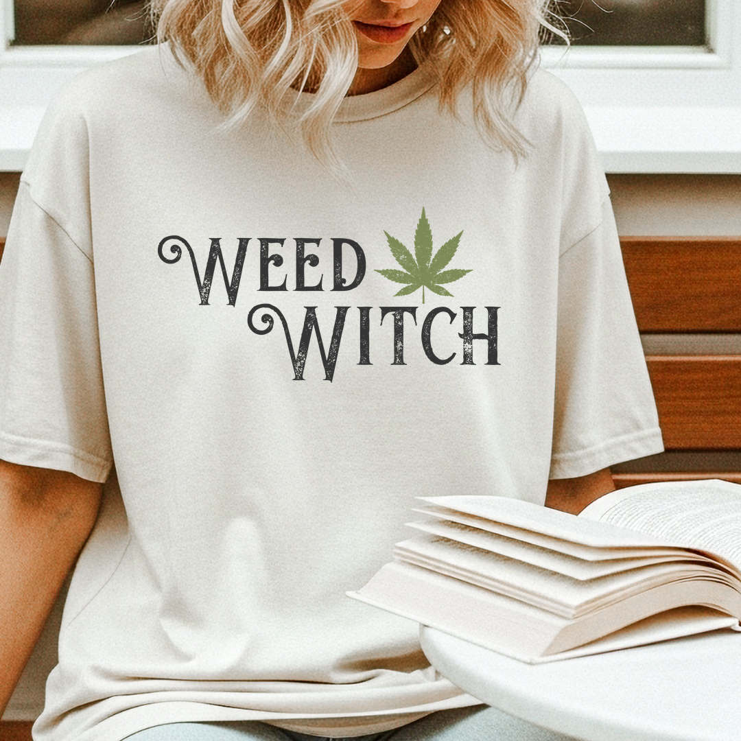 A woman wearing a cream-colored t-shirt with a 'WEED WITCH' graphic featuring a green cannabis leaf, sitting next to an open book.