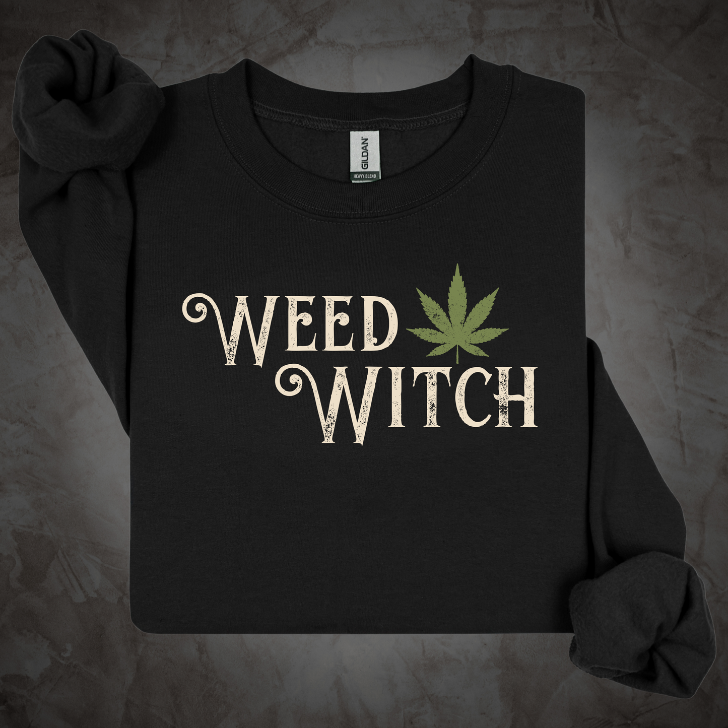 A black long-sleeve t-shirt with 'WEED WITCH' printed in a distressed cream font next to a green cannabis leaf graphic, displayed on a textured gray background.