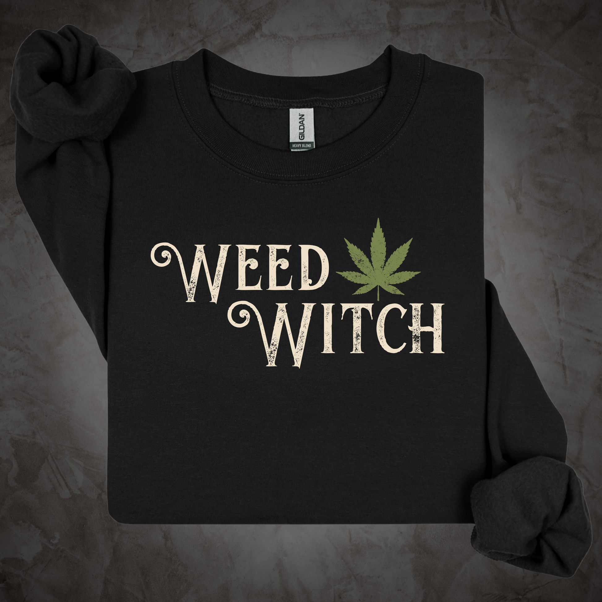 A black long-sleeve t-shirt with 'WEED WITCH' printed in a distressed cream font next to a green cannabis leaf graphic, displayed on a textured gray background.