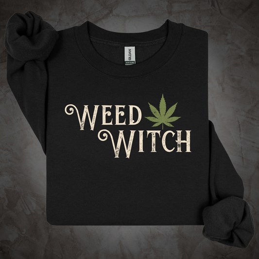 A black long-sleeve t-shirt with 'WEED WITCH' printed in a distressed cream font next to a green cannabis leaf graphic, displayed on a textured gray background.