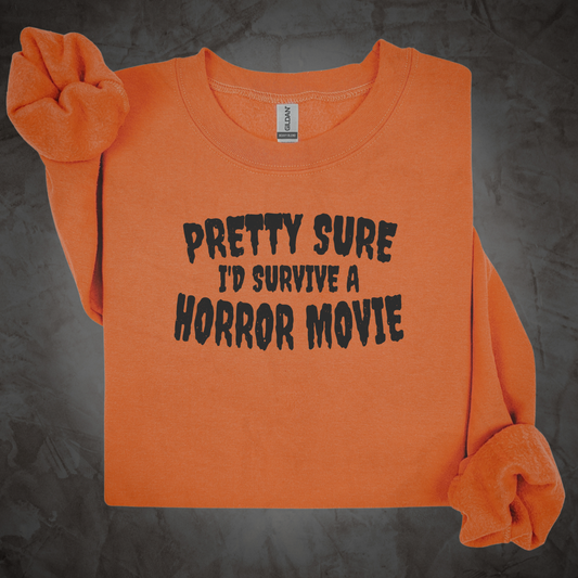 An orange long-sleeve sweatshirt with the text 'PRETTY SURE I'D SURVIVE A HORROR MOVIE' printed in black across the chest, styled with rolled cuffs and a matching orange scrunchie on a dark textured background.