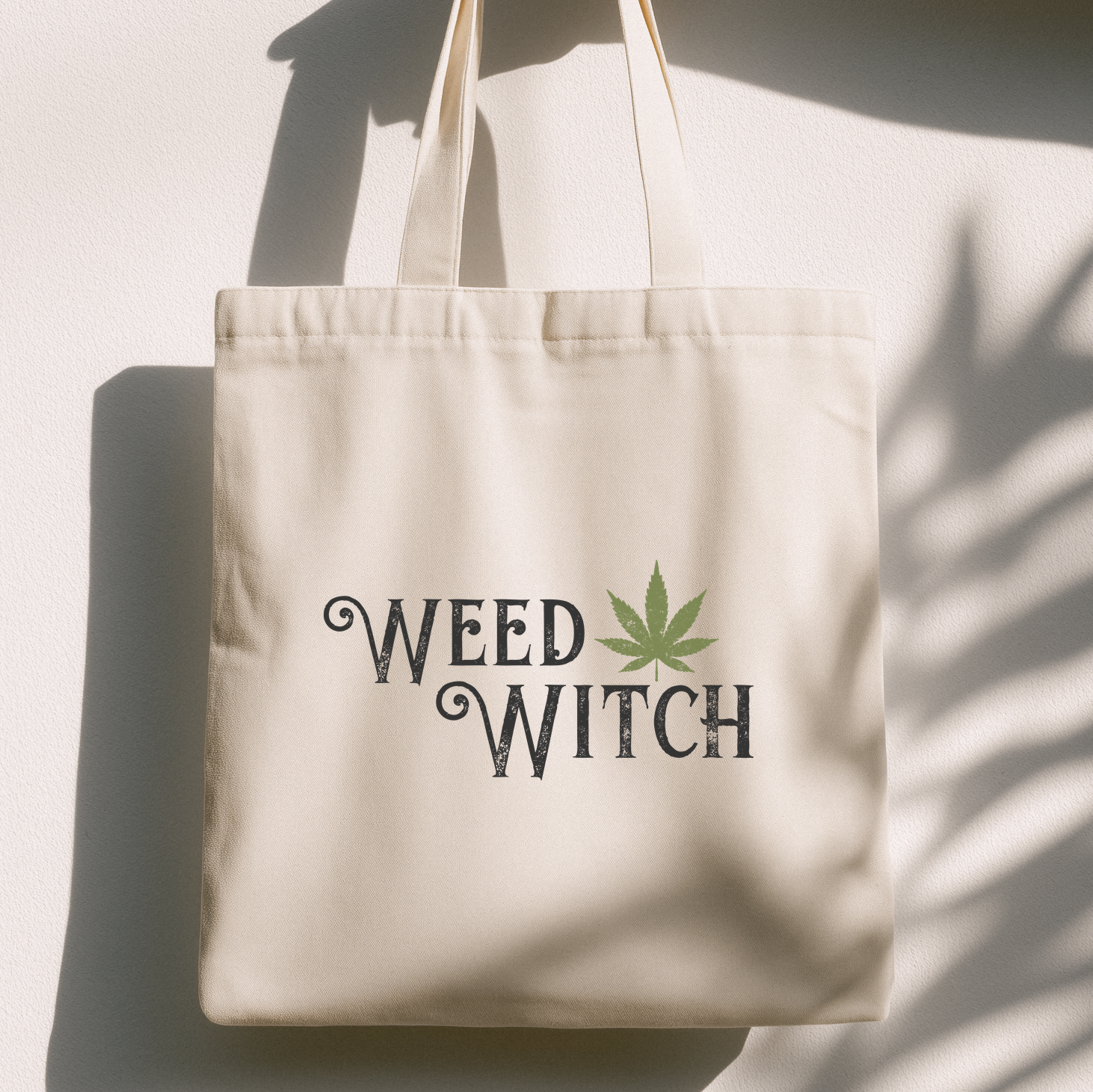 A cream-colored canvas tote bag with the words 'WEED WITCH' printed in a dark gray gothic font, accented by a green cannabis leaf. The bag is photographed hanging against a white wall with dramatic shadows cast across it.