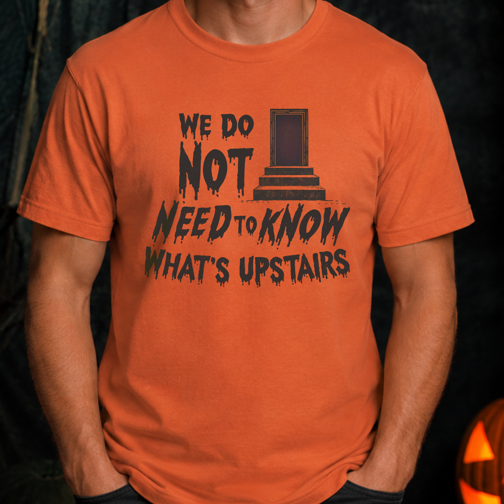 An orange short-sleeve graphic t-shirt worn by a model. The shirt features a black distressed print that reads 'WE DO NOT NEED TO KNOW WHAT'S UPSTAIRS' alongside a simple illustration of a tombstone.