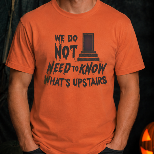 An orange short-sleeve graphic t-shirt worn by a model. The shirt features a black distressed print that reads 'WE DO NOT NEED TO KNOW WHAT'S UPSTAIRS' alongside a simple illustration of a tombstone.