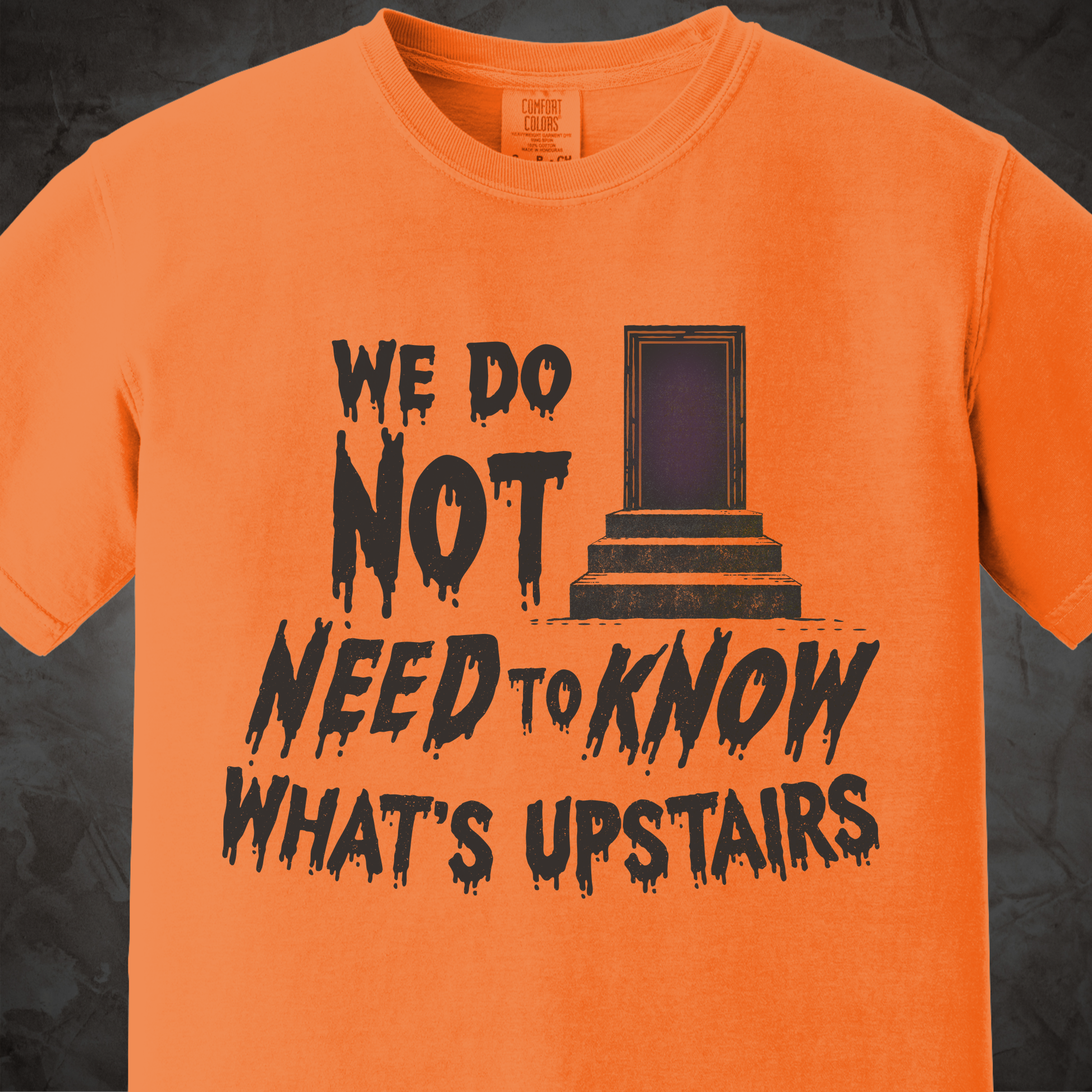 An orange short-sleeve crewneck t-shirt featuring a black distressed graphic that reads 'WE DO NOT NEED TO KNOW WHAT'S UPSTAIRS' alongside a dark purple door illustration, displayed against a dark textured background.