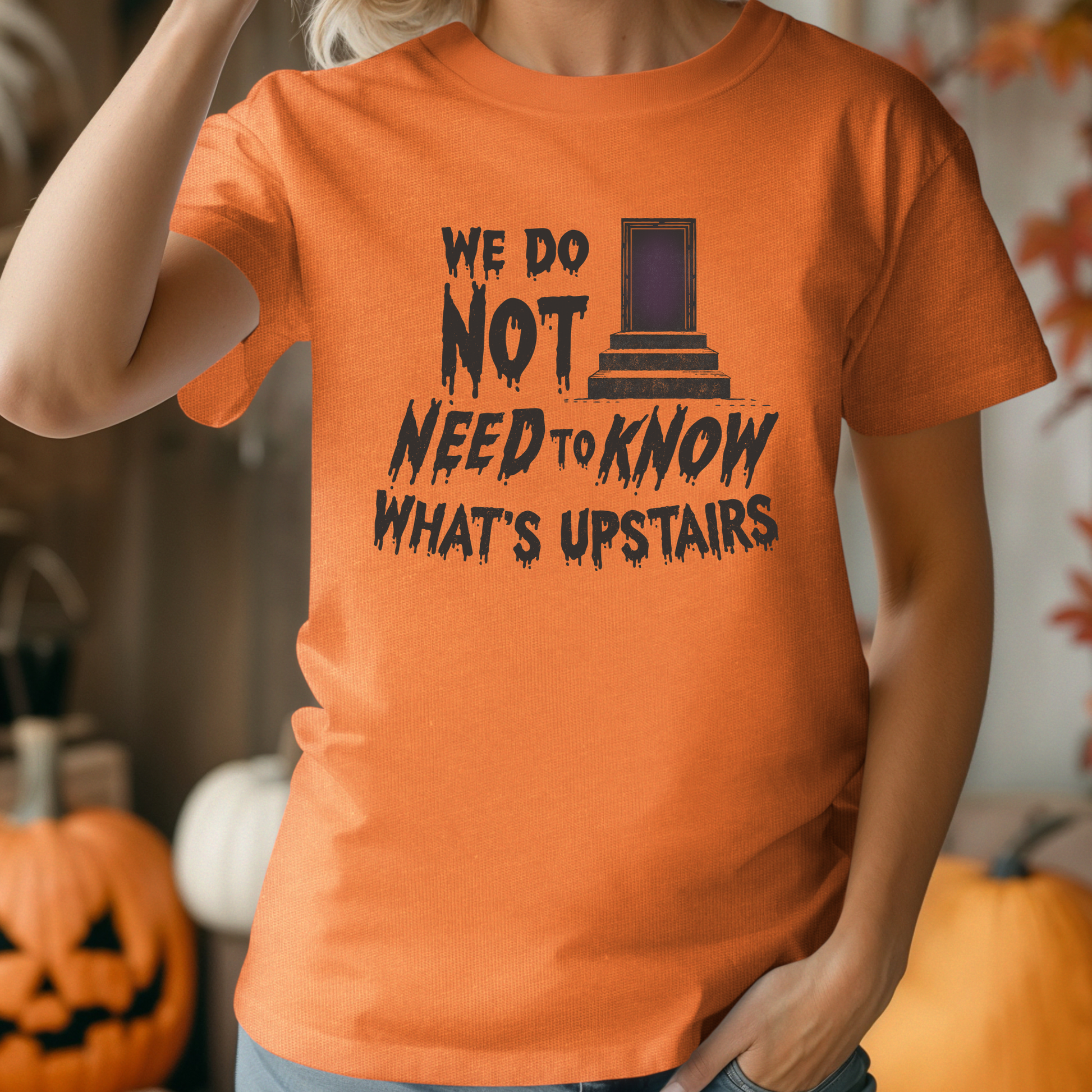 An orange short-sleeve graphic T-shirt worn by a person in a Halloween-themed setting with pumpkins in the background. The shirt features a black printed design with the text 'WE DO NOT NEED TO KNOW WHAT'S UPSTAIRS' and a tombstone graphic.