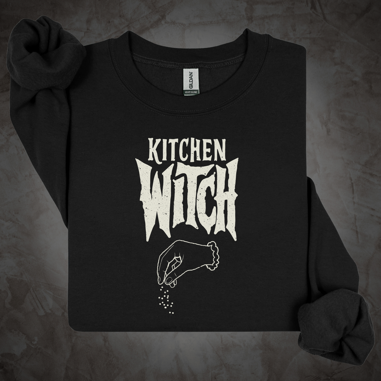 A black long-sleeve crewneck sweatshirt with a distressed white graphic reading 'KITCHEN WITCH' and a hand sprinkling spices, laid flat on a textured gray background.