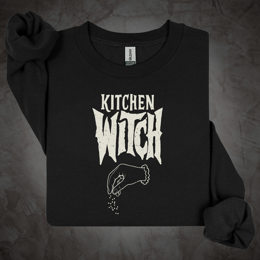 A black long-sleeve crewneck sweatshirt with a distressed white graphic reading 'KITCHEN WITCH' and a hand sprinkling spices, laid flat on a textured gray background.