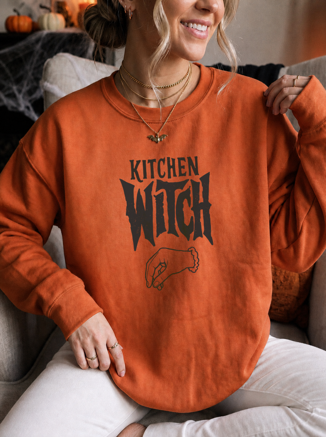 A woman wearing an orange sweatshirt with a black graphic that reads 'KITCHEN WITCH' and a small witch hat illustration, sitting on a couch in a cozy indoor setting.