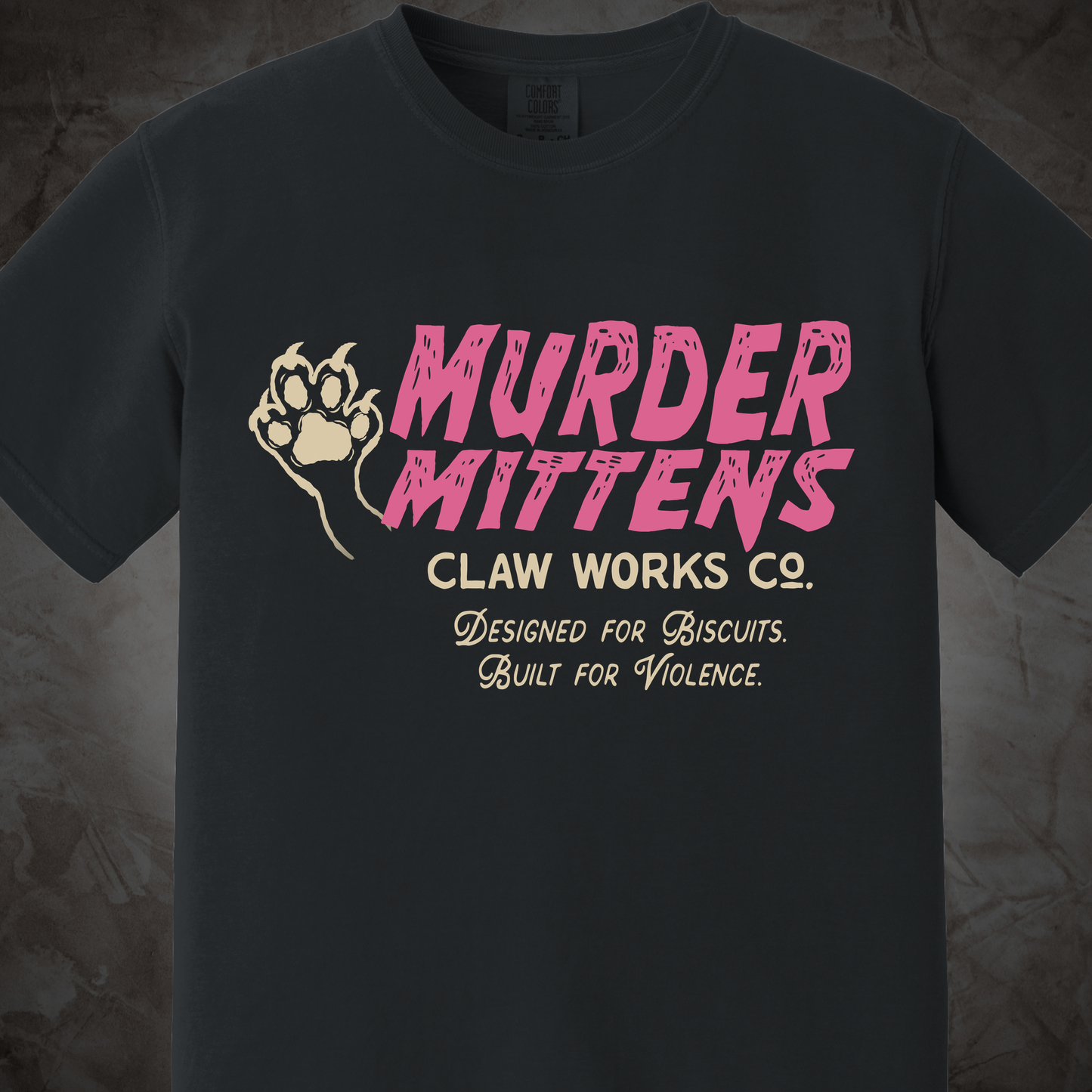 A dark charcoal gray t-shirt featuring a graphic print with the text 'MURDER MITTENS' in pink, 'CLAW WORKS CO.' in beige, and 'Designed for Biscuits.