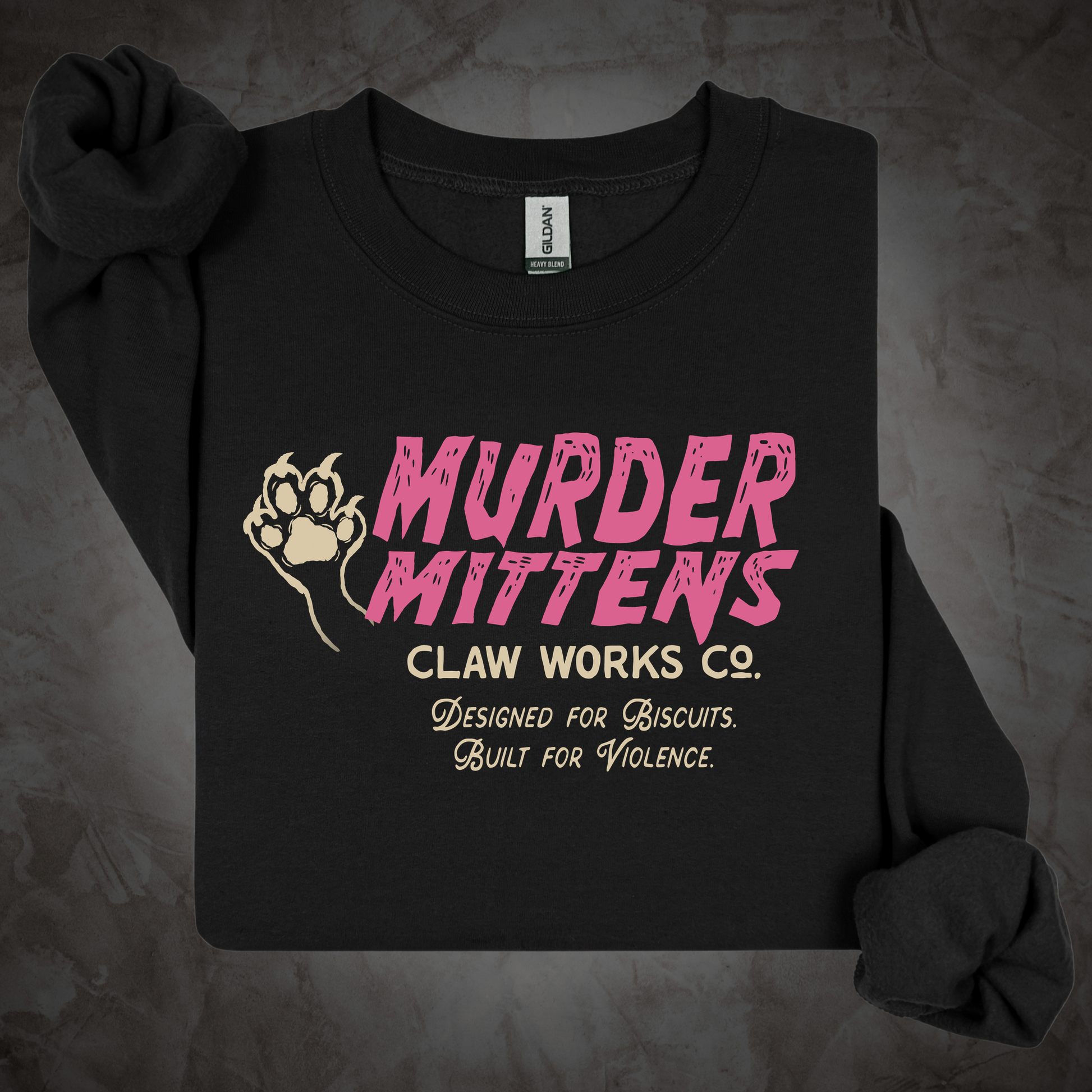 A black long-sleeve crewneck sweatshirt laid flat on a textured gray background. The front features a graphic print with a beige paw print and the text 'MURDER MITTENS CLAW WORKS CO.