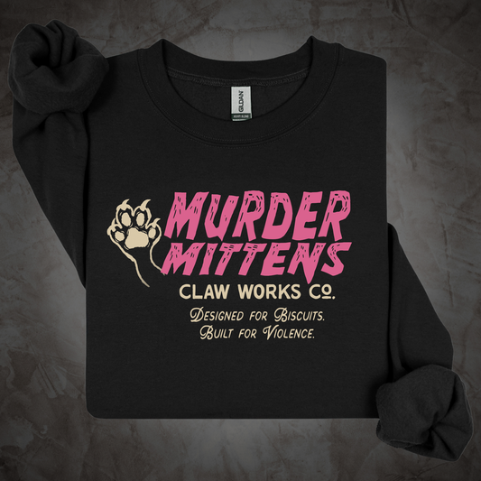 A black long-sleeve crewneck sweatshirt laid flat on a textured gray background. The front features a graphic print with a beige paw print and the text 'MURDER MITTENS CLAW WORKS CO.