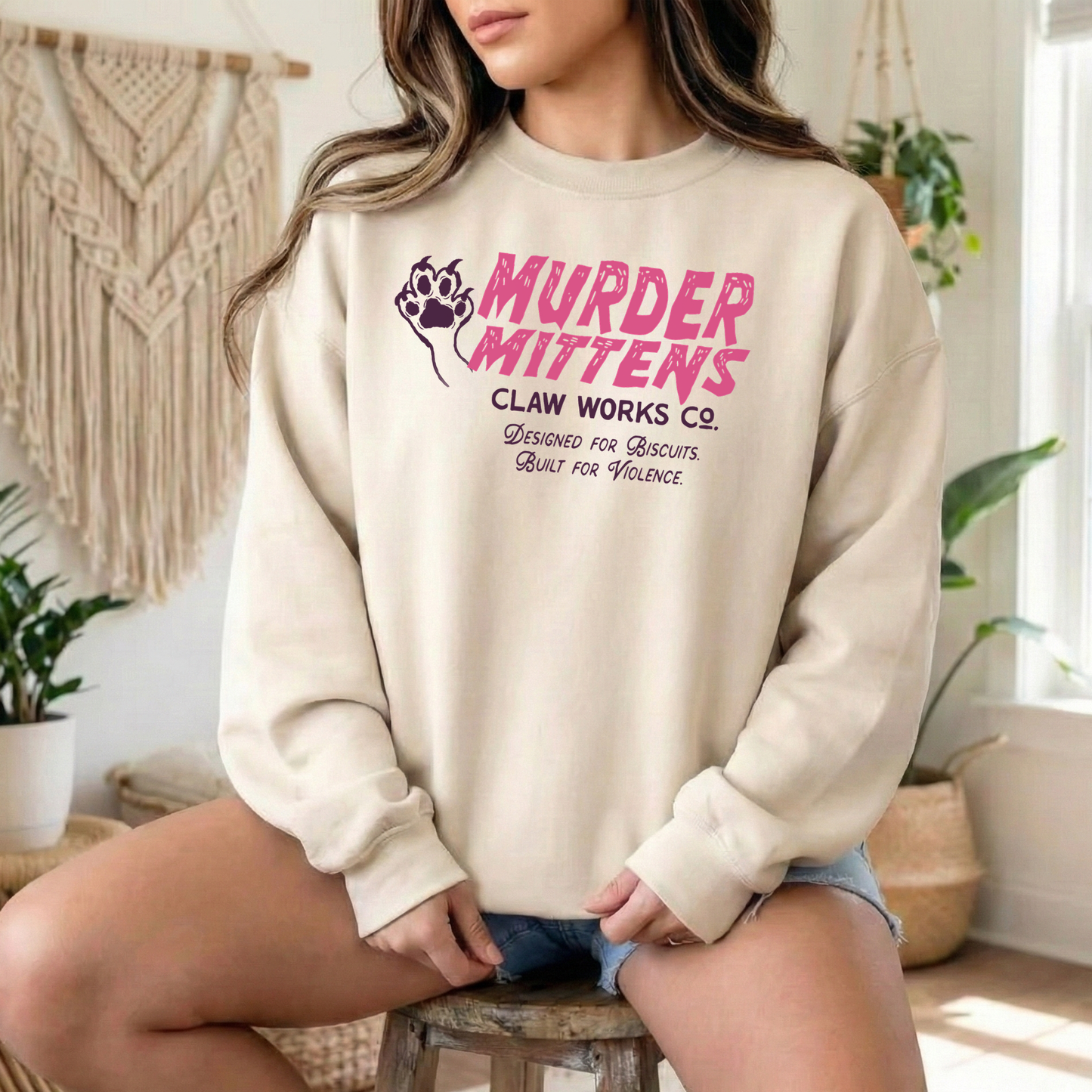 A woman wearing a cream-colored sweatshirt with a pink and purple graphic that reads 'MURDER MITTENS CLAW WORKS CO.' while sitting on a stool in a bright, boho-style room.