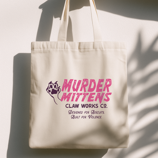 A cream canvas tote bag with pink and purple text reading 'MURDER MITTENS CLAW WORKS CO.' and a paw print graphic, hanging against a white wall with strong shadow patterns.