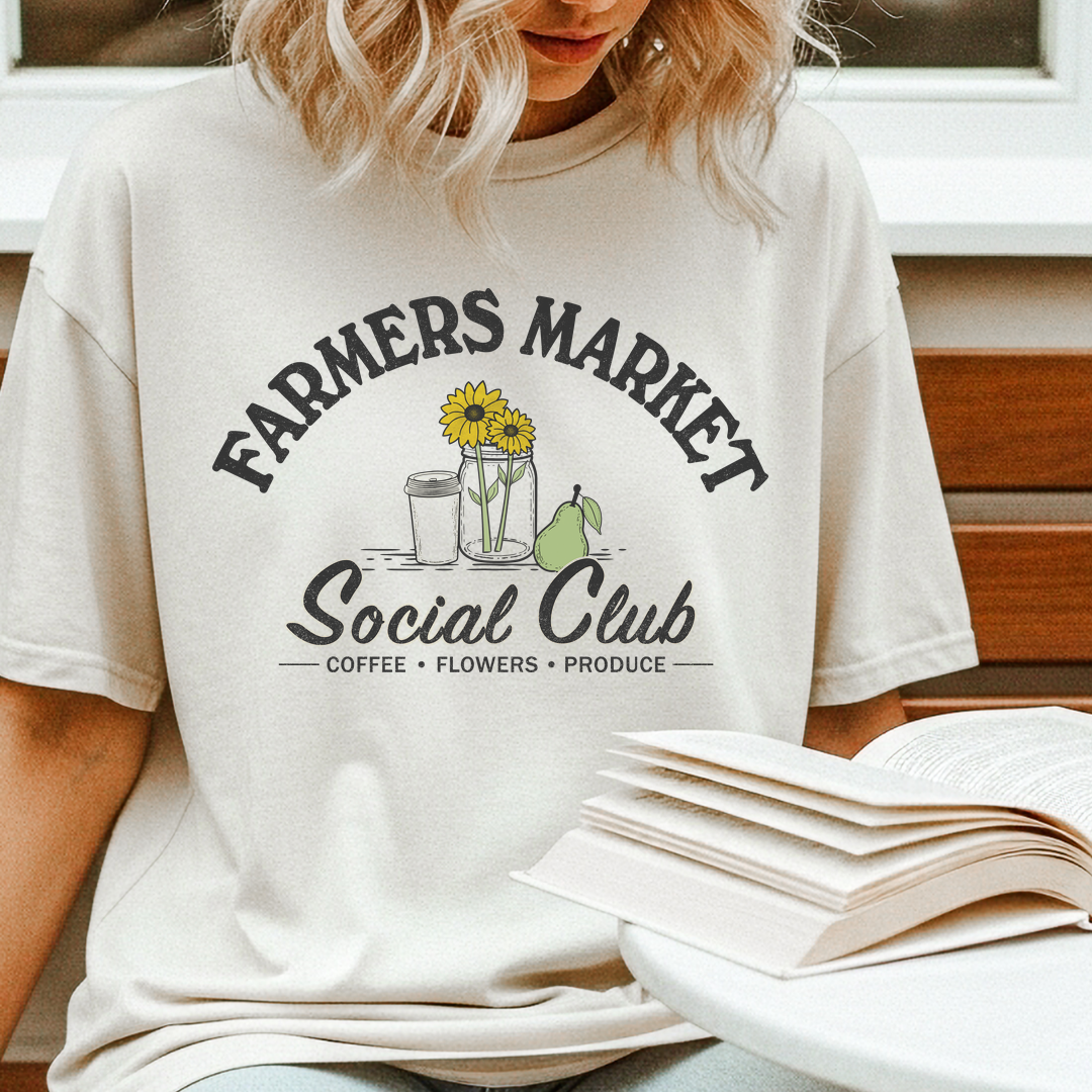 A woman wearing a cream-colored graphic t-shirt with 'FARMERS MARKET Social Club' printed in black, featuring a sunflower, coffee cup, and pear illustration. She is sitting on a wooden bench with an open book in the foreground.