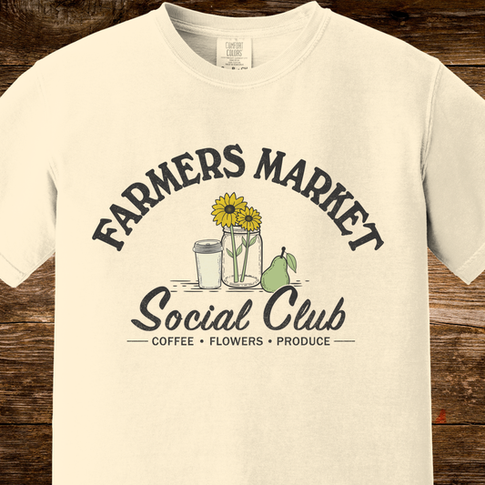 An off-white short-sleeve t-shirt with a graphic print reading 'FARMERS MARKET Social Club' and 'COFFEE • FLOWERS • PRODUCE' around an illustration of sunflowers, a cup, a jar, and a pear, displayed on a rustic dark wood background.