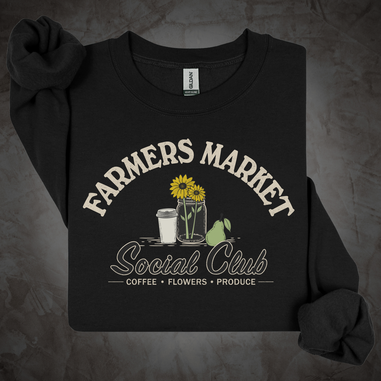 A cream-colored long-sleeve t-shirt with rolled cuffs, featuring a graphic print that reads 'FARMERS MARKET Social Club' with illustrations of sunflowers, a coffee cup, a pear, and a jar. The shirt is laid flat on a dark wood plank background.