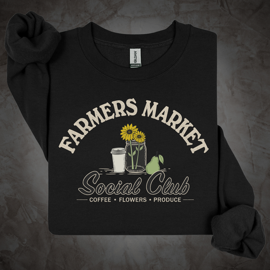 A cream-colored long-sleeve t-shirt with rolled cuffs, featuring a graphic print that reads 'FARMERS MARKET Social Club' with illustrations of sunflowers, a coffee cup, a pear, and a jar. The shirt is laid flat on a dark wood plank background.