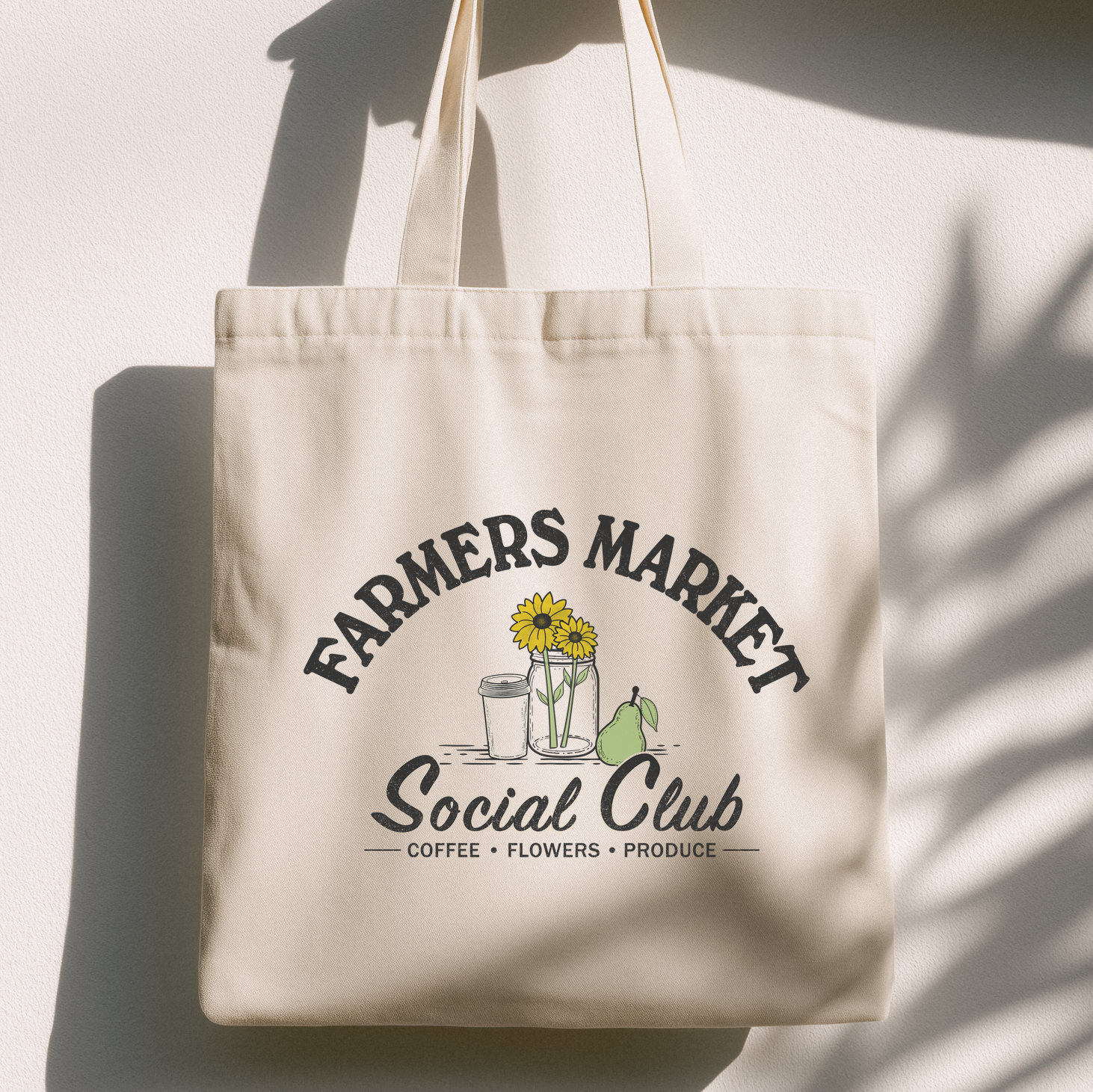 A cream canvas tote bag with a printed graphic reading 'FARMERS MARKET Social Club' and small illustrations of sunflowers, a coffee cup, and a pear, hanging against a white wall with strong shadow patterns.