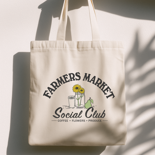 A cream canvas tote bag with a printed graphic reading 'FARMERS MARKET Social Club' and small illustrations of sunflowers, a coffee cup, and a pear, hanging against a white wall with strong shadow patterns.