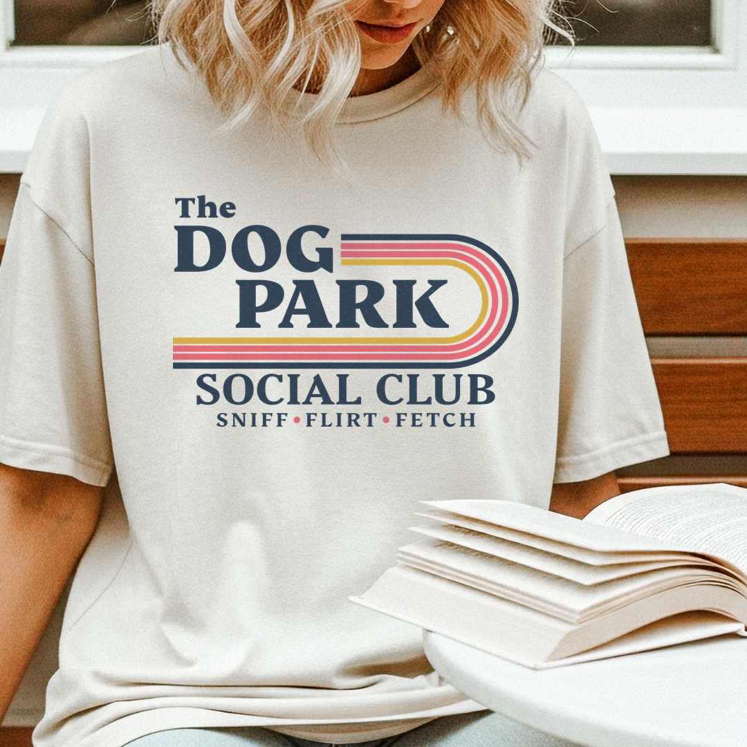 A woman wearing a cream-colored graphic t-shirt with a retro-style 'The Dog Park Social Club' print in navy and pink, sitting on a wooden bench next to an open book.